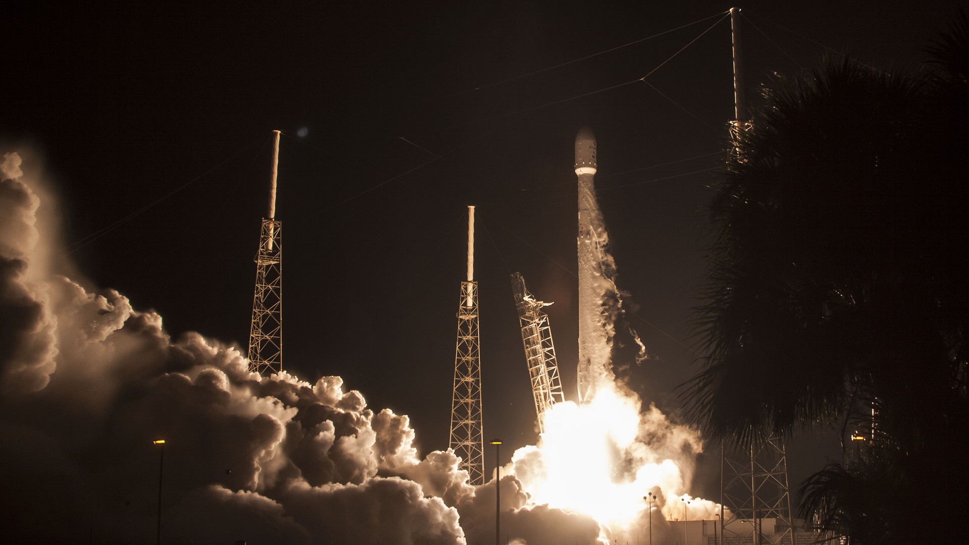 A SpaceX Falcon 9 lifting off at night from a Florida launch pad, engines flaring, exhaust plume lit against a dark sky.