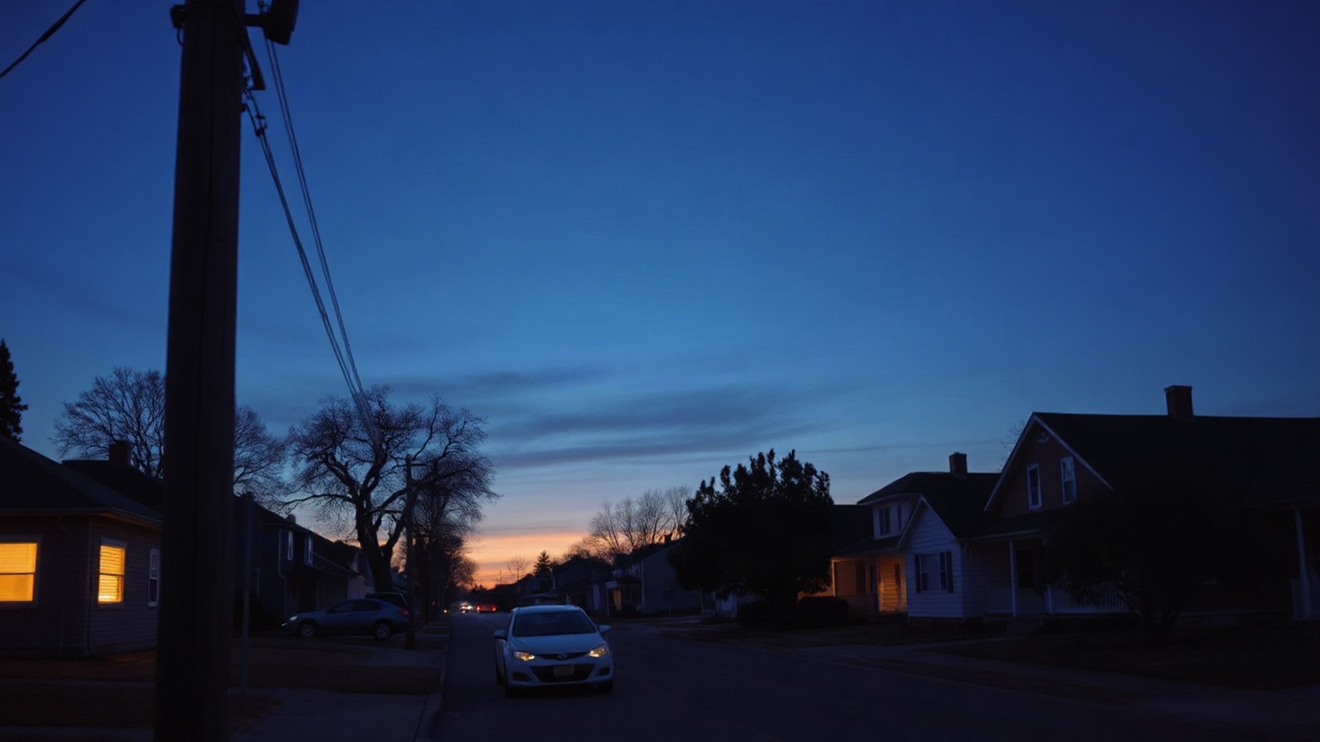 A suburban residential street at dusk, lit windows glowing behind a wooden utility pole and its distribution lines. The same grid that AI data centers lean on runs the neighborhood, and regulators are still deciding who pays for the load it carries. Photo: The Signal / AI-generated.